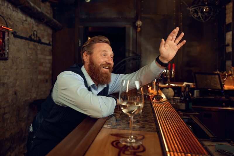 Lelaki mabuk tersenyum gembira sambil melambai tangan sambil duduk di kaunter bar